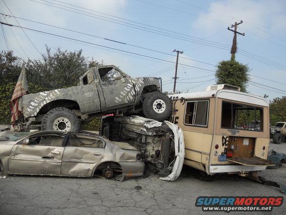 Mud-splattered pickup truck on top of a crushed sedan and a beige bus