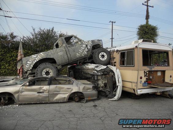 Wrecked pickup truck on top of sedan, next to a bus. Tire tracks painted on truck