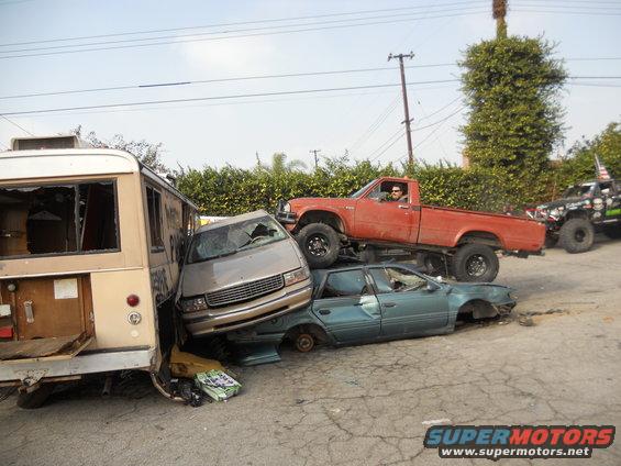 Orange pickup truck stacked on a teal sedan, with a bus and minivan in foreground