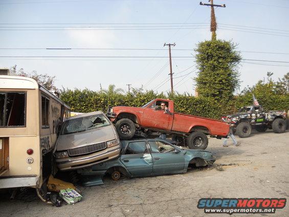 red pickup truck on top of sedan, with van and another truck nearby