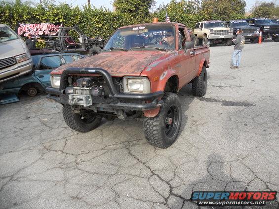 Red pickup truck with a winch, lifted suspension, and off-road tires. The body is rusty
