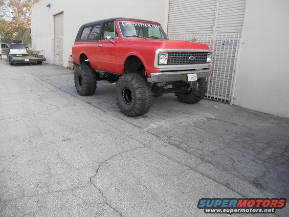 red lifted SUV with big tires, black top, and 'DESMADRE' banner on windshield. Another vehicle on a trailer is parked behind it