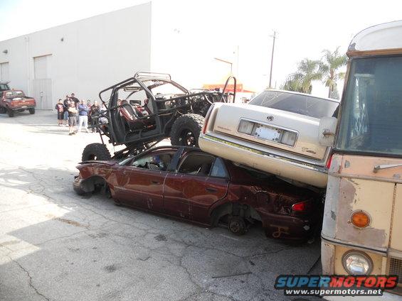 Several wrecked cars are stacked, with a custom off-road vehicle on top. A bus is parked to the right