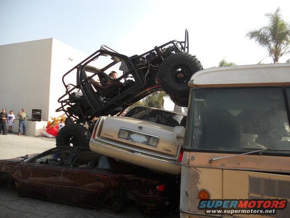 Off-road buggy atop a Cadillac and a sedan, next to a bus