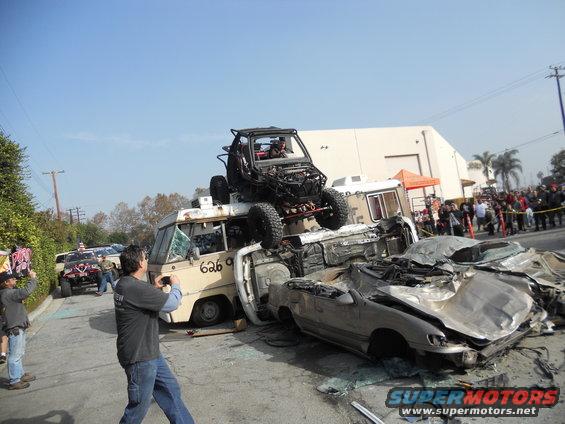Off-road buggy atop a pile of wrecked cars and a van, with spectators watching