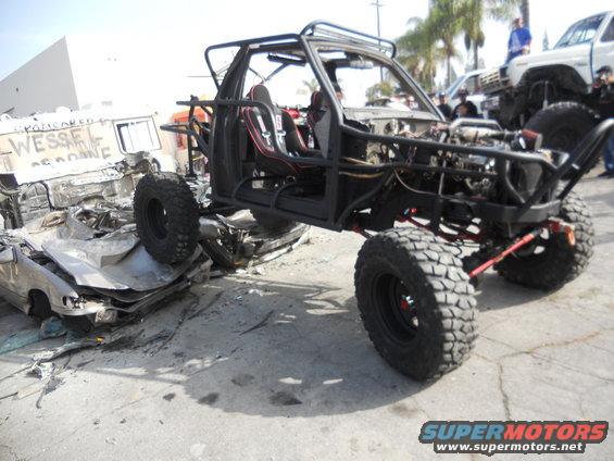 Custom off-road buggy with roll cage, racing seats, and large tires, parked next to a wrecked car and a white pickup truck