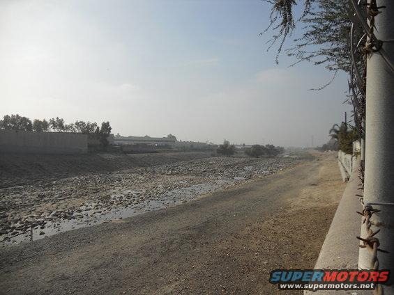 Dry riverbed with a dirt road and concrete barrier, barbed wire fence, hazy sky