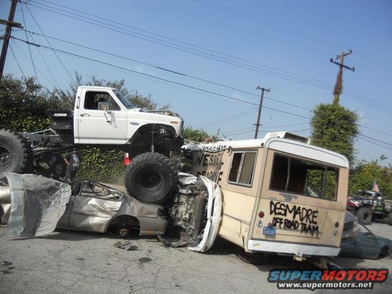 White pickup truck with large tires is on top of a wrecked bus and car. Text on bus reads 'DESMADRE OFF ROAD TEAM'