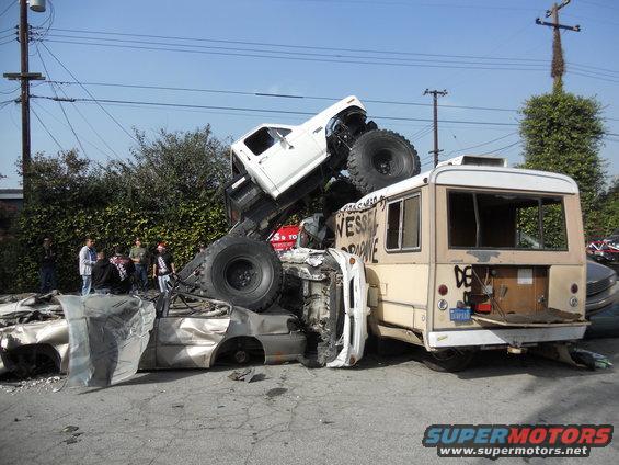 Monster truck on top of a wrecked bus and car, with people watching