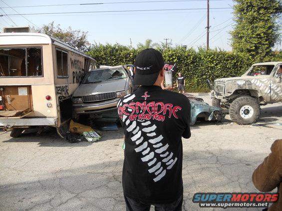 Person in a 'DESMADRE OFFROAD TEAM' shirt looks at a scene with a wrecked bus, a damaged sedan, and a large off-road truck