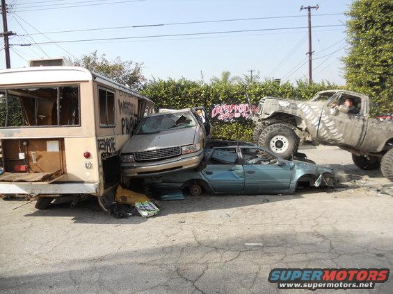 Wrecked cars and a bus piled up, with a lifted monster truck on top