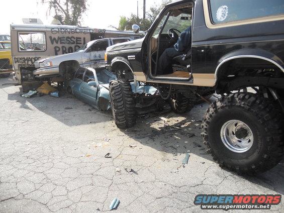 1986 Ford Bronco parked over a crushed sedan, with a van and another car in the background