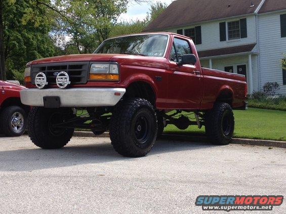 1996 Ford F-150 pickup truck, lifted with large tires, parked in front of a house. Another red vehicle is partially visible on the left