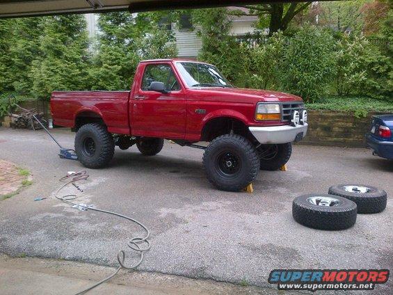 Red 1996 Ford F-150 pickup truck with lifted suspension and off-road tires, parked in a driveway with a jack and spare tires nearby