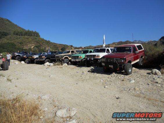 Red and white 1987 Ford Bronco, 3/4 front view, parked off-road with other trucks