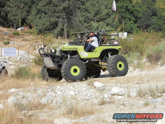 Modified green jeep with large tires and roll cage driving on a dirt path