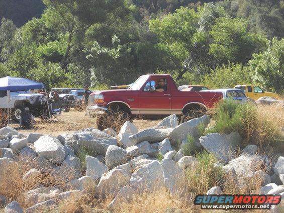 Red Bronco with lifted suspension parked near rocks, driver visible. Other vehicles and a tent are in the background