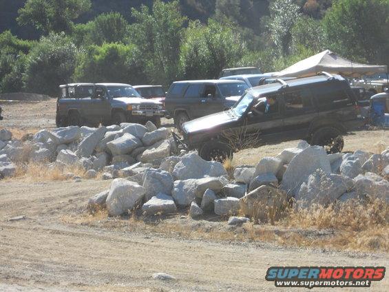 Dark SUV with roof rack driving over rocks, other SUVs and trucks in background
