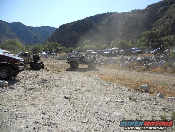 Several off-road vehicles parked and driving in a dusty, rocky canyon with mountains in the background