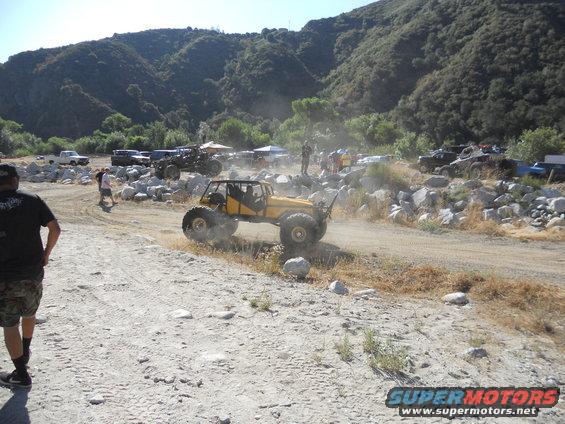 Yellow off-road buggy with large tires driving on a dirt path, kicking up dust. Mountains in background