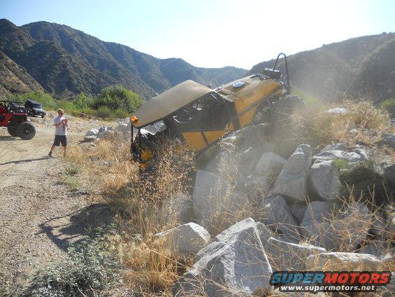 Yellow off-road vehicle tilted on rocks with a person standing nearby and another vehicle in the background