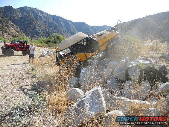 Yellow off-road vehicle tipped on its side in rocks with a red Jeep nearby