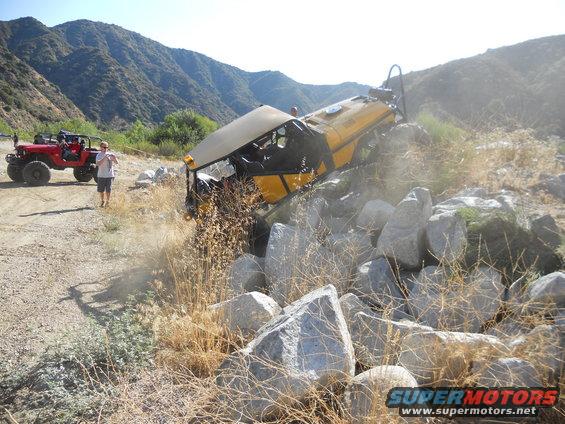 Yellow off-road vehicle tilted on a pile of rocks, with a red Jeep and a person standing nearby on a dirt path