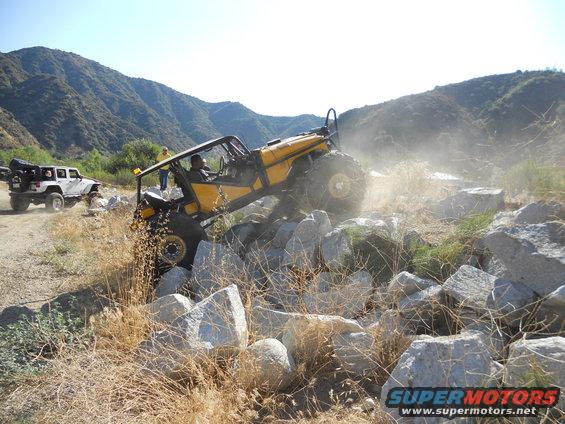 Yellow off-road vehicle navigating a rocky terrain with a white Jeep in the background
