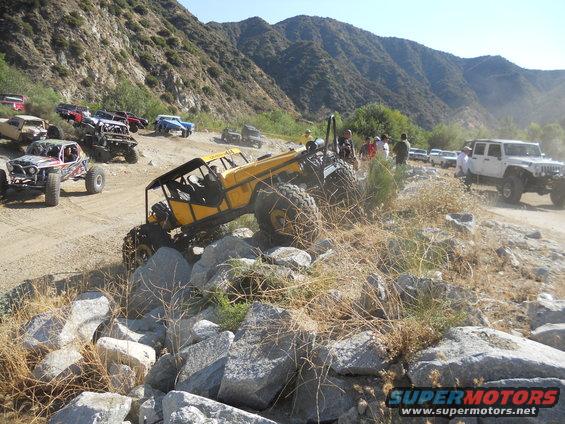 Several off-road vehicles, including a yellow buggy and a white Jeep, navigate a rocky mountain trail