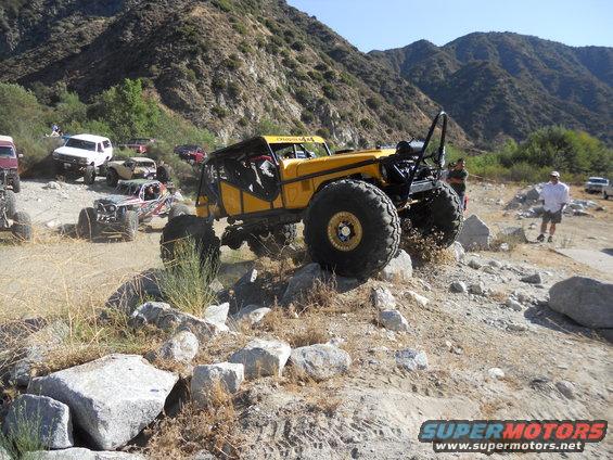 Yellow custom buggy with huge tires crawling over rocks, other off-road vehicles and people around