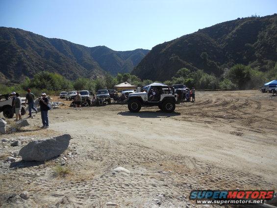 Several off-road vehicles parked in a dirt lot with mountains in the background. A white Jeep is in the foreground