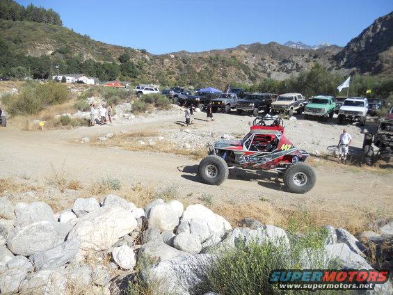 Red off-road buggy with number 442 on the side, parked on a dirt road with other vehicles in the background