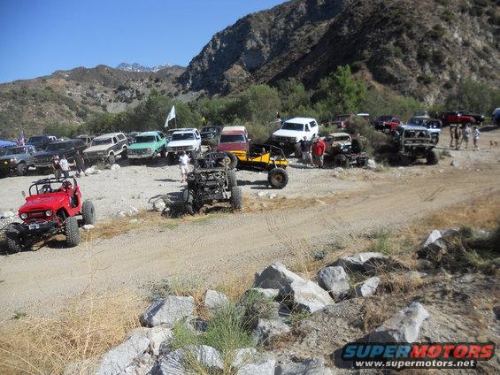 Red Jeep-like off-road vehicle in foreground, with many other off-road vehicles gathered in a dry, rocky, mountainous area