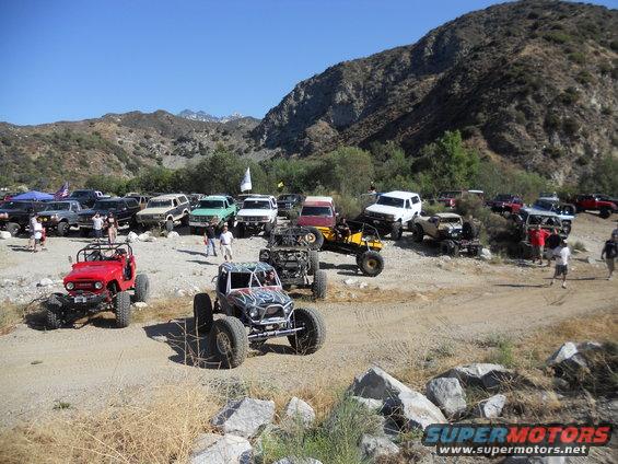 Many off-road vehicles parked in a dusty canyon with mountains in the background