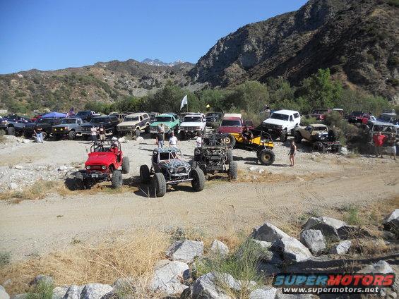 Many off-road vehicles parked in a rocky, mountainous area under a clear blue sky