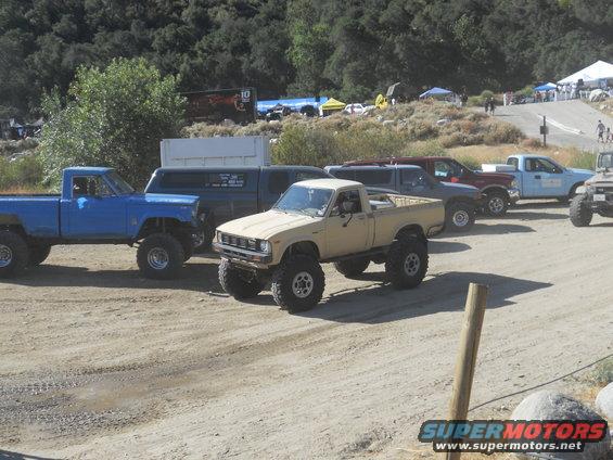 Several lifted pickup trucks parked on a dirt lot with trees and tents in the background