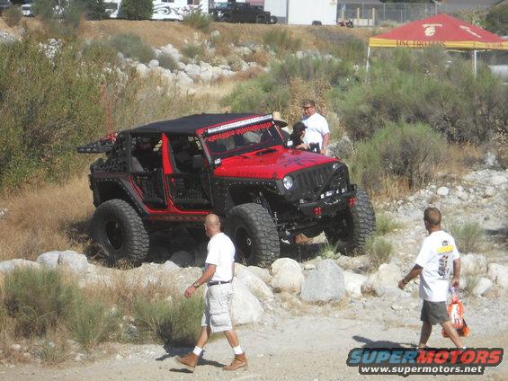 Red Jeep Wrangler Rubicon with off-road tires and lights, parked on rocky terrain with people around