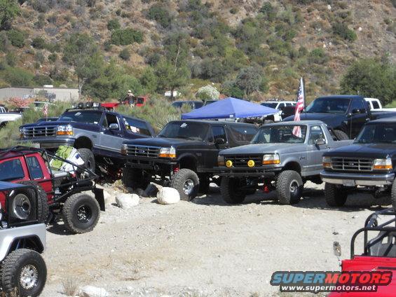 Several lifted Ford Bronco and F-Series trucks parked on dirt with hills in background. One truck has an American flag
