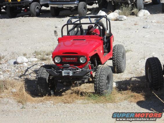 Red Toyota Land Cruiser FJ40 with roll cage and large tires parked on dirt, other vehicles in background