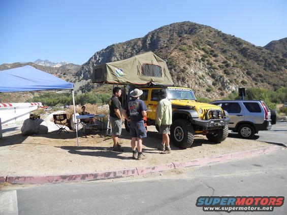 Yellow Jeep Wrangler with rooftop tent and silver Honda CR-V parked on dirt lot