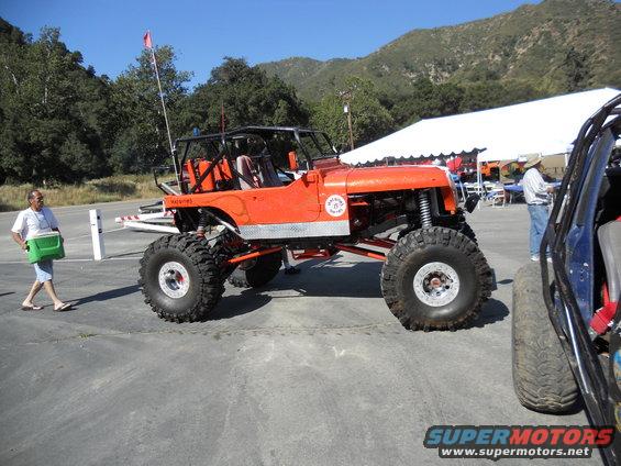 Orange off-road vehicle with large tires and roll cage, parked next to another vehicle