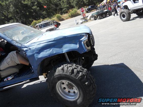 Blue off-road truck with large tires, missing front end. Other vehicles and people in background