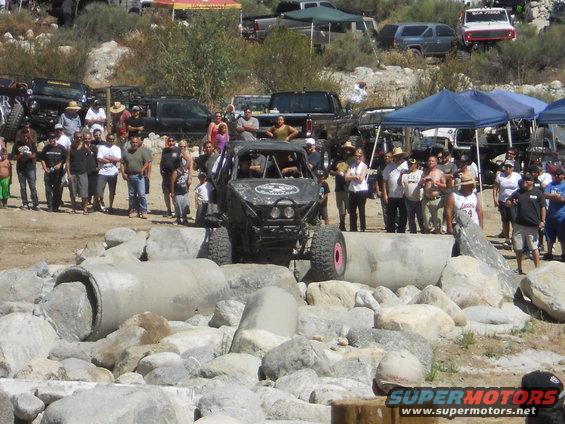Off-road buggy on rocks, crowd watching. Other trucks and tents in background