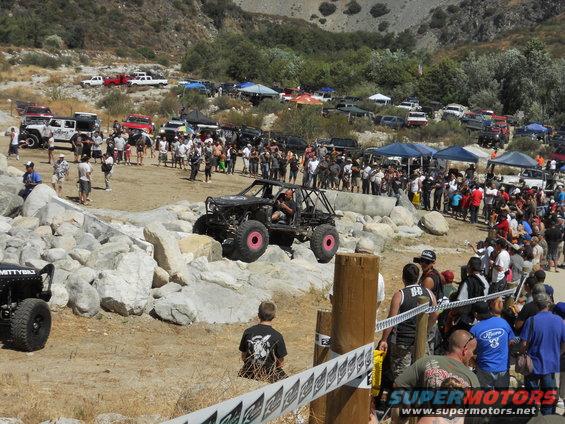 Off-road buggy with pink wheels on rocky terrain, crowd watching