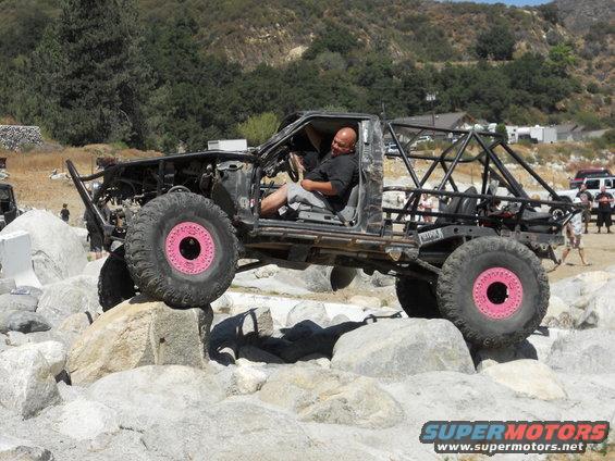 Custom rock crawler with pink wheels on rocks, driver inside. Other vehicles and people in background