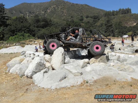 Off-road buggy with pink wheels climbing over large rocks on a sunny day. Driver is visible inside
