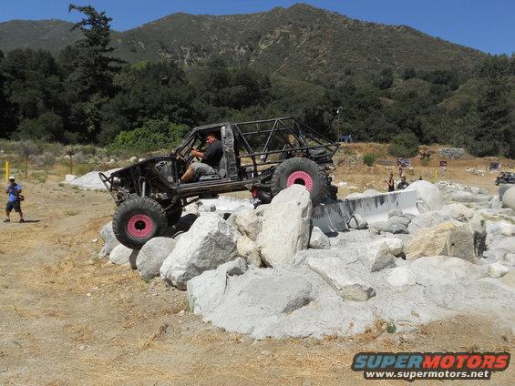 Modified off-road vehicle with roll cage navigating rocks, pink wheel accents visible