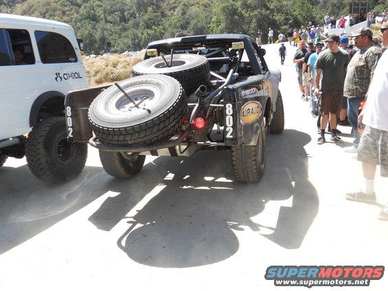 Off-road race truck with two spare tires mounted on the back, next to a white SUV