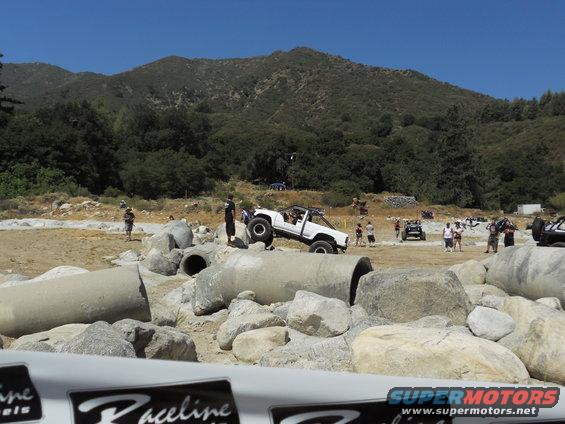 White off-road truck climbing over large concrete pipes and rocks, with other vehicles and people around