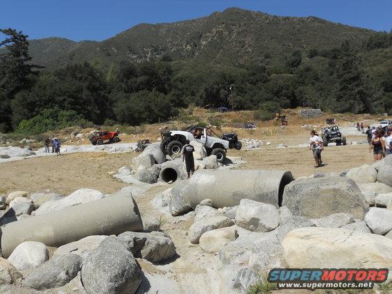 Several off-road vehicles, including a white Jeep-like vehicle and an orange buggy, are on a dirt course with large rocks and concrete pipes